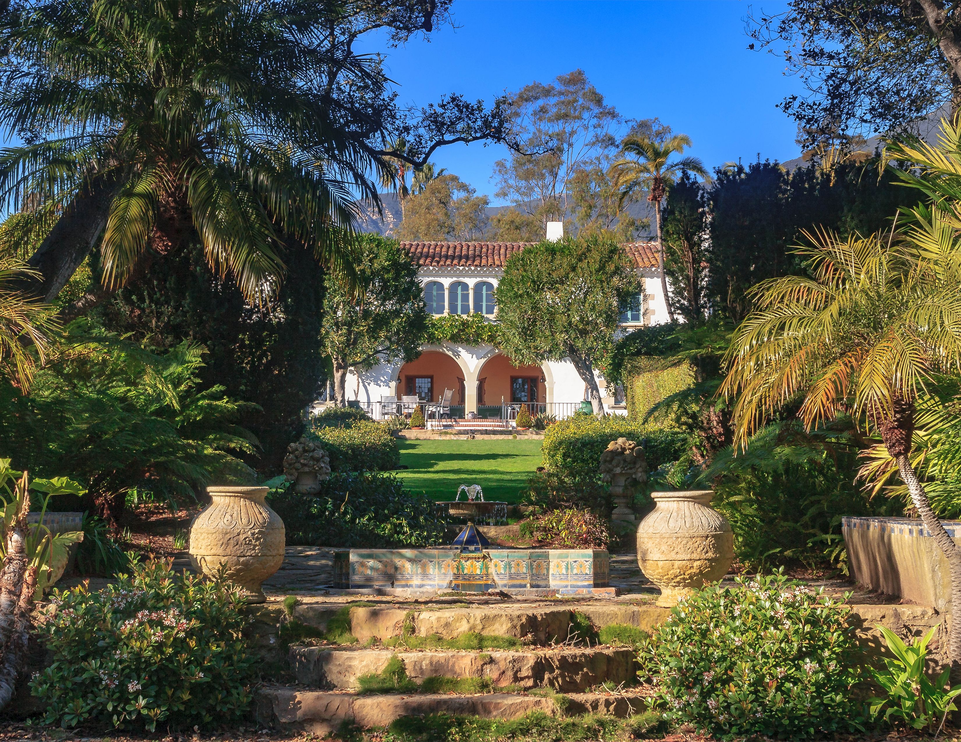 Stone steps and a fountain on the grounds of Casa de Herrero, with the historic home framed with plants, trees and a blue sky.